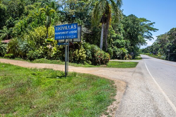 Un letrero de Tikivillas Rainforest Lodge con un camino de tierra que conduce a una carretera pavimentada.