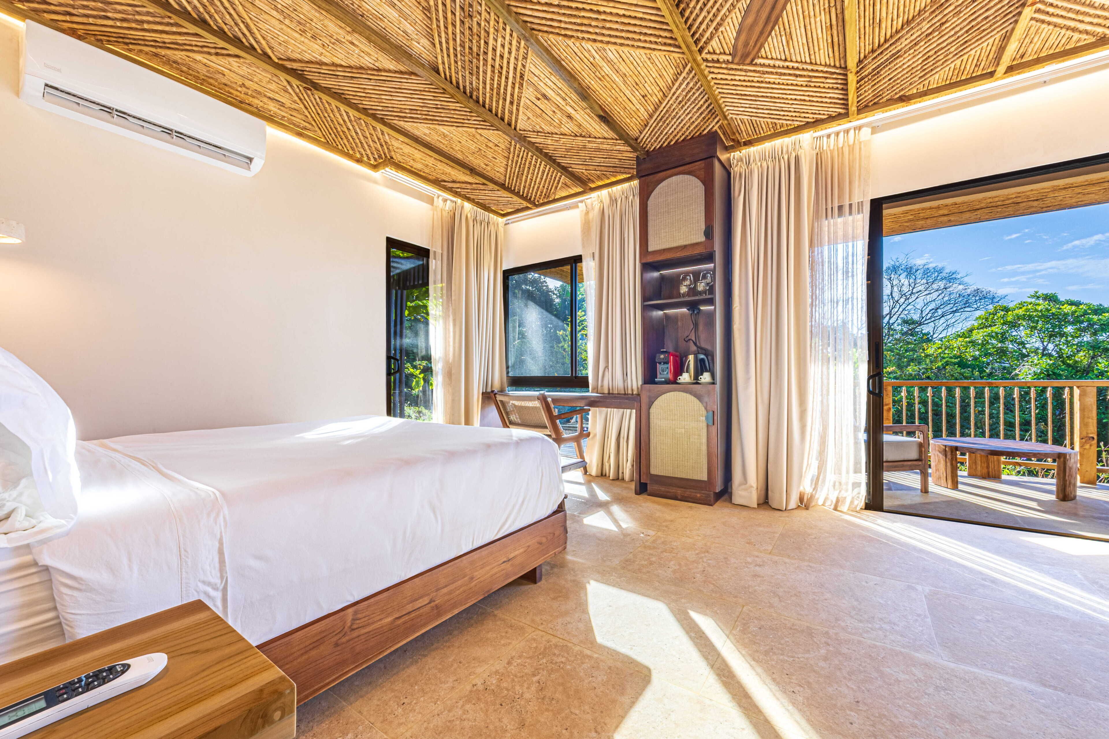 A bedroom with a bed, a wooden desk, a chair, a TV, and a balcony with a view of greenery.
