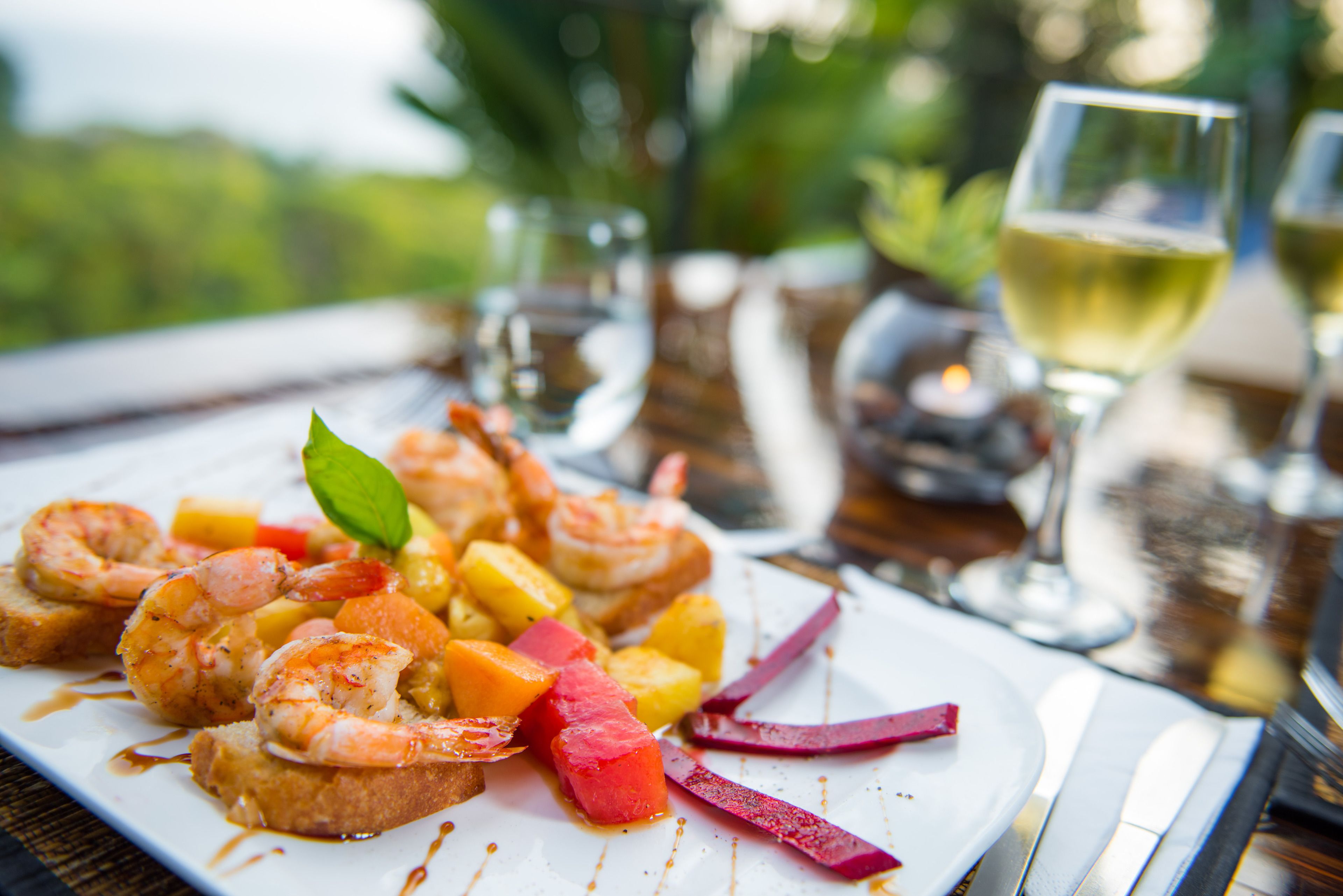 Un plato de camarones y fruta sobre una mesa, con una copa de vino blanco y una vela al fondo.