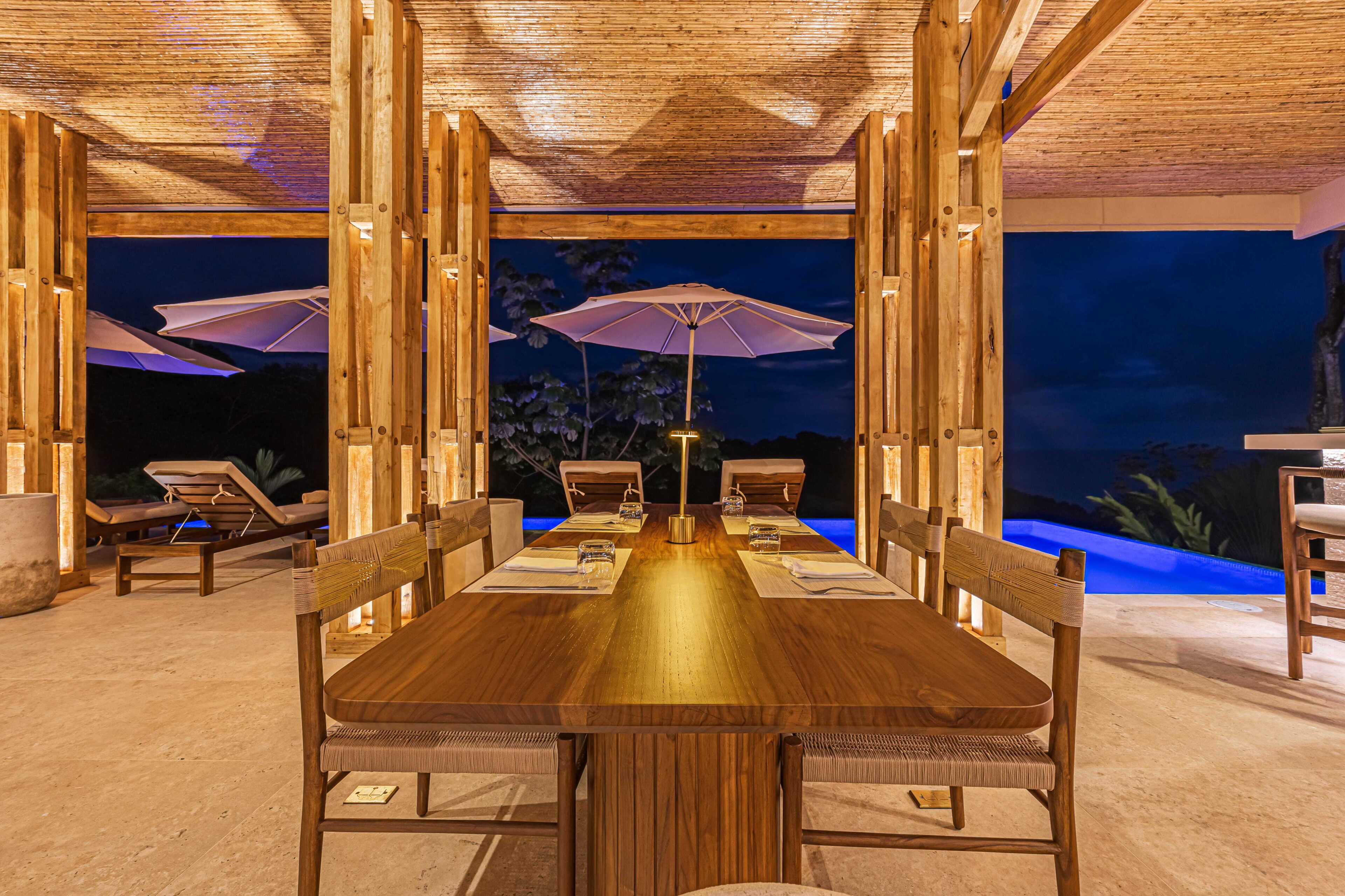 A wooden dining table with chairs, umbrellas, and a pool view at night.