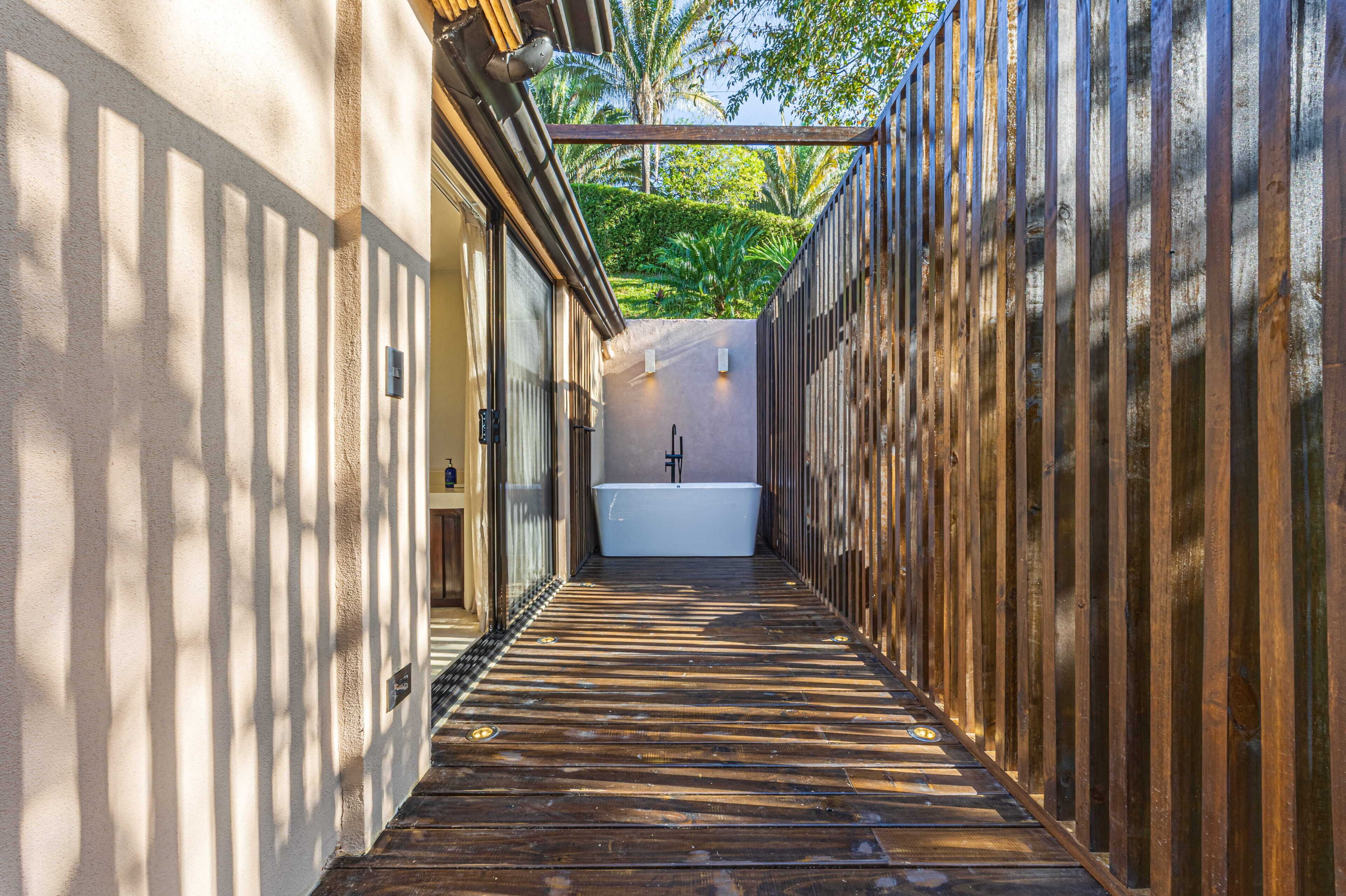 A narrow pathway leading to a white bathtub in a spa-like setting with wooden railings and a view of greenery.