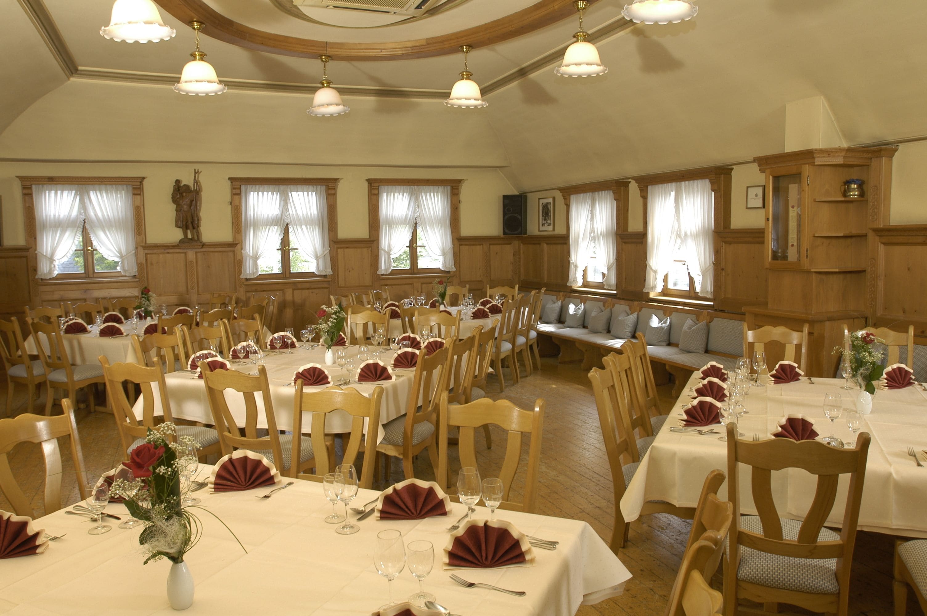 Ein großer Speisesaal mit mehreren gedeckten Tischen für eine festliche Veranstaltung, mit Holzsesseln und weißen Tischdecken.