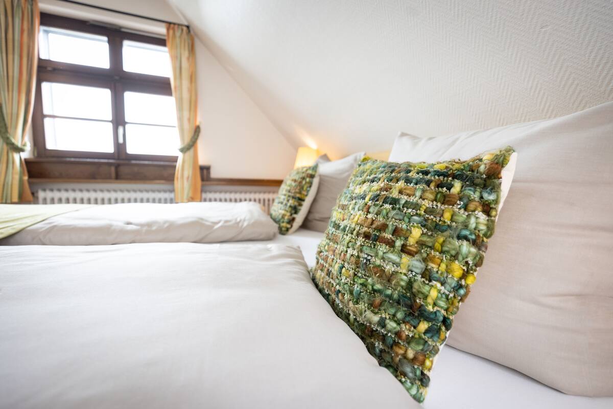 A neatly made bed with white linens and a colorful, textured pillow.