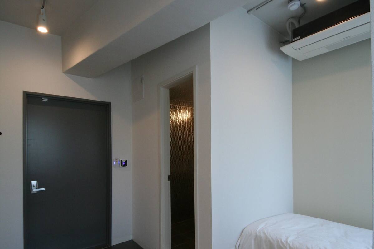 A modern bedroom with a single bed, a small white dresser, a door, and a pair of shoes on the floor.