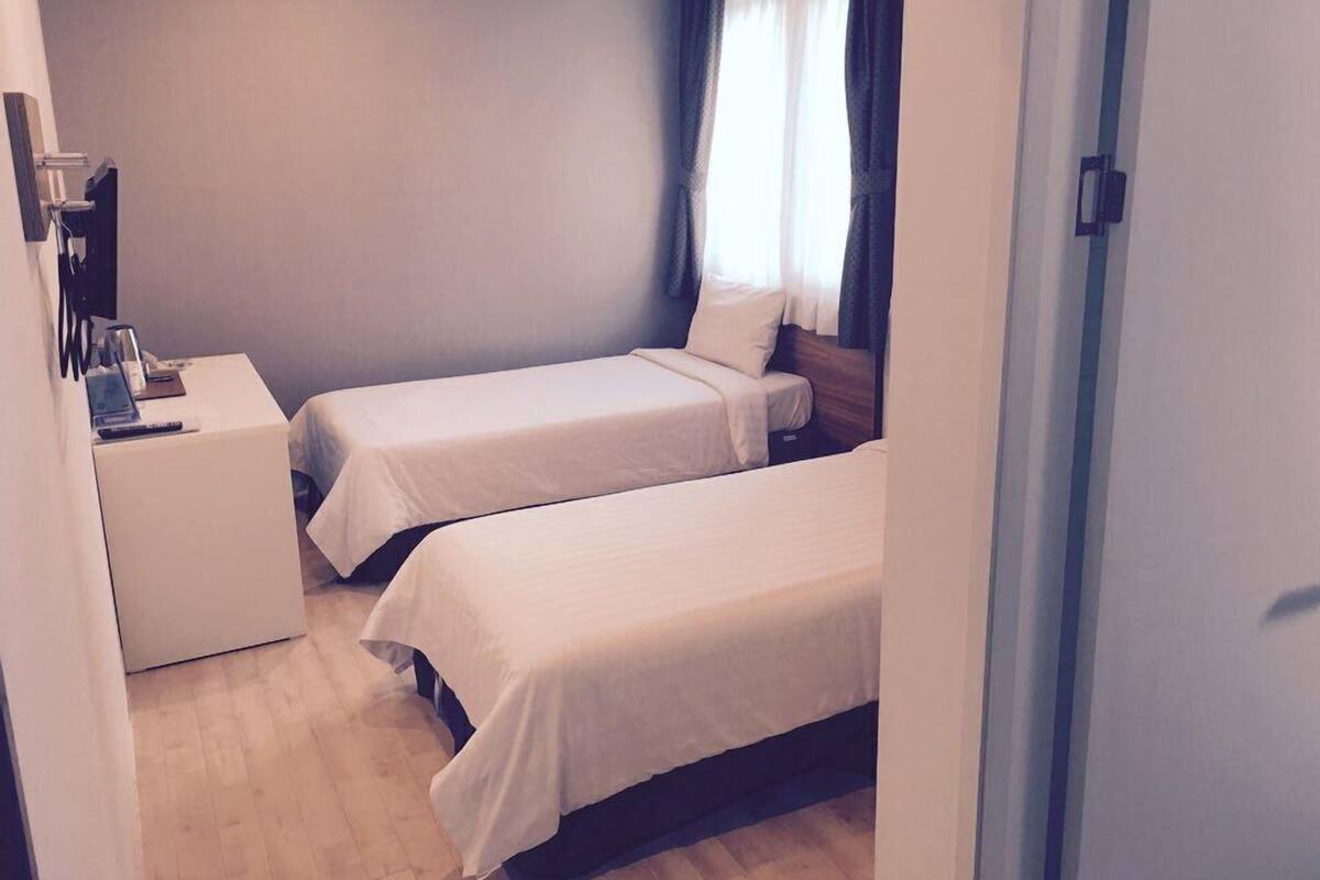 A small, clean room with two single beds, a bedside table, and a window with curtains.