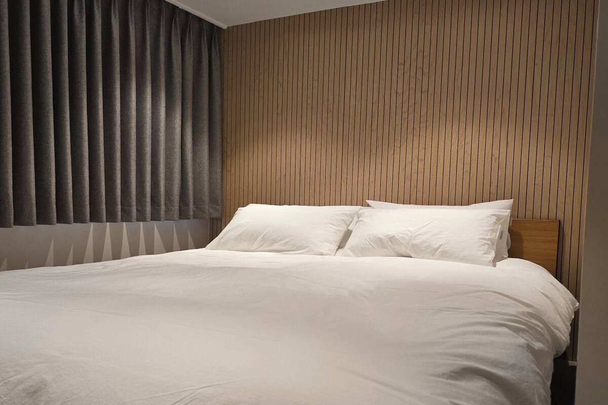 A modern bedroom with a large bed, wooden panel wall, and a window with dark curtains.