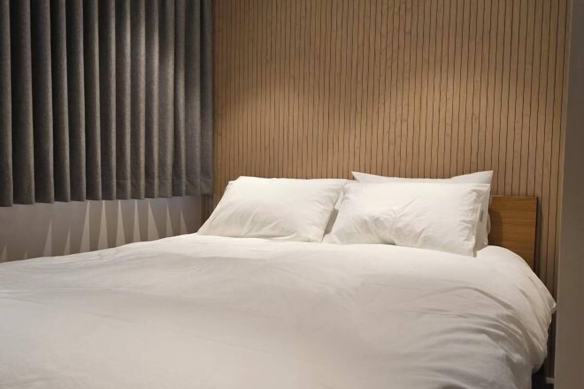 A bed with white bedding, a wooden headboard, and vertical blinds on the window.