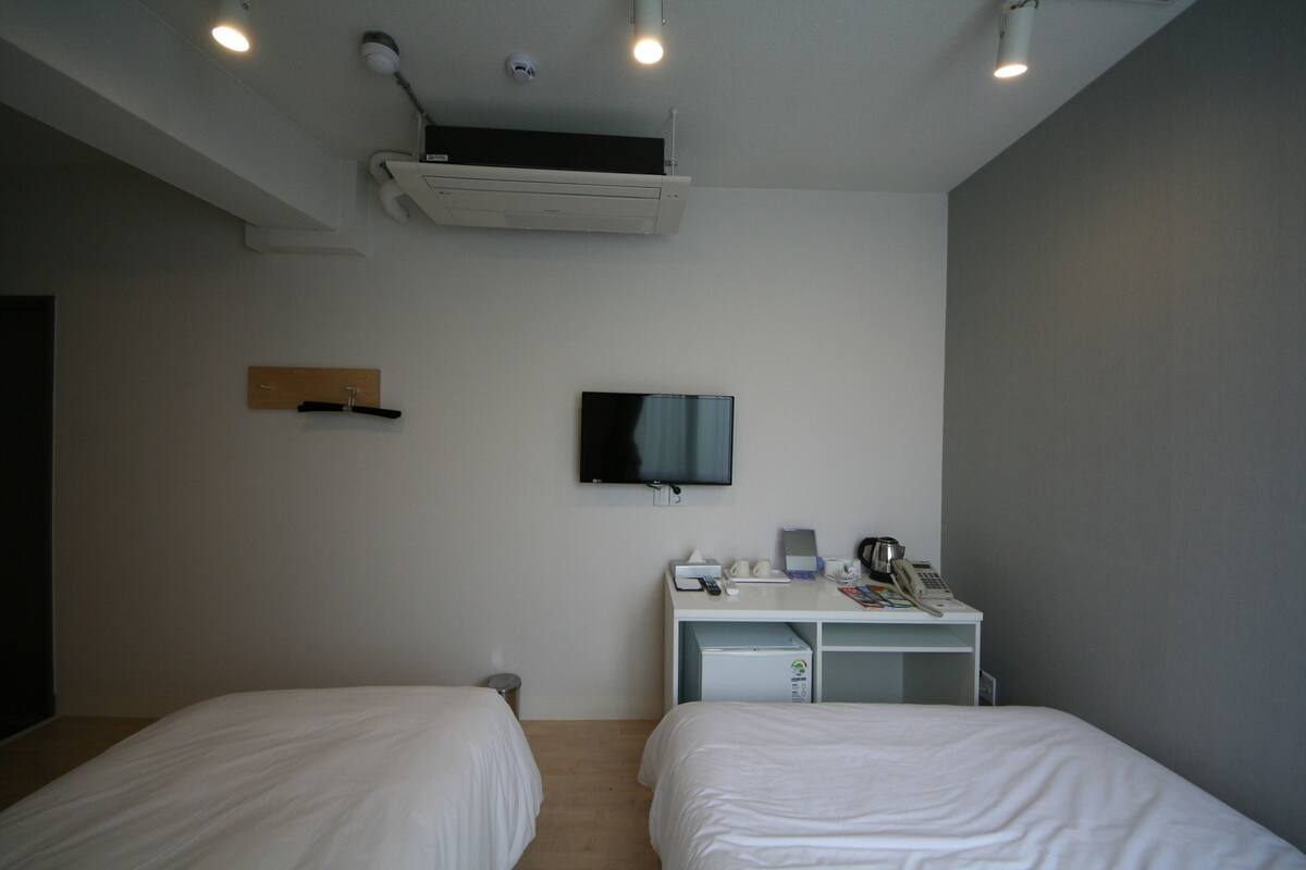 A hotel room with two beds, a desk with a flat-screen TV, a small fridge, and a trash bin.