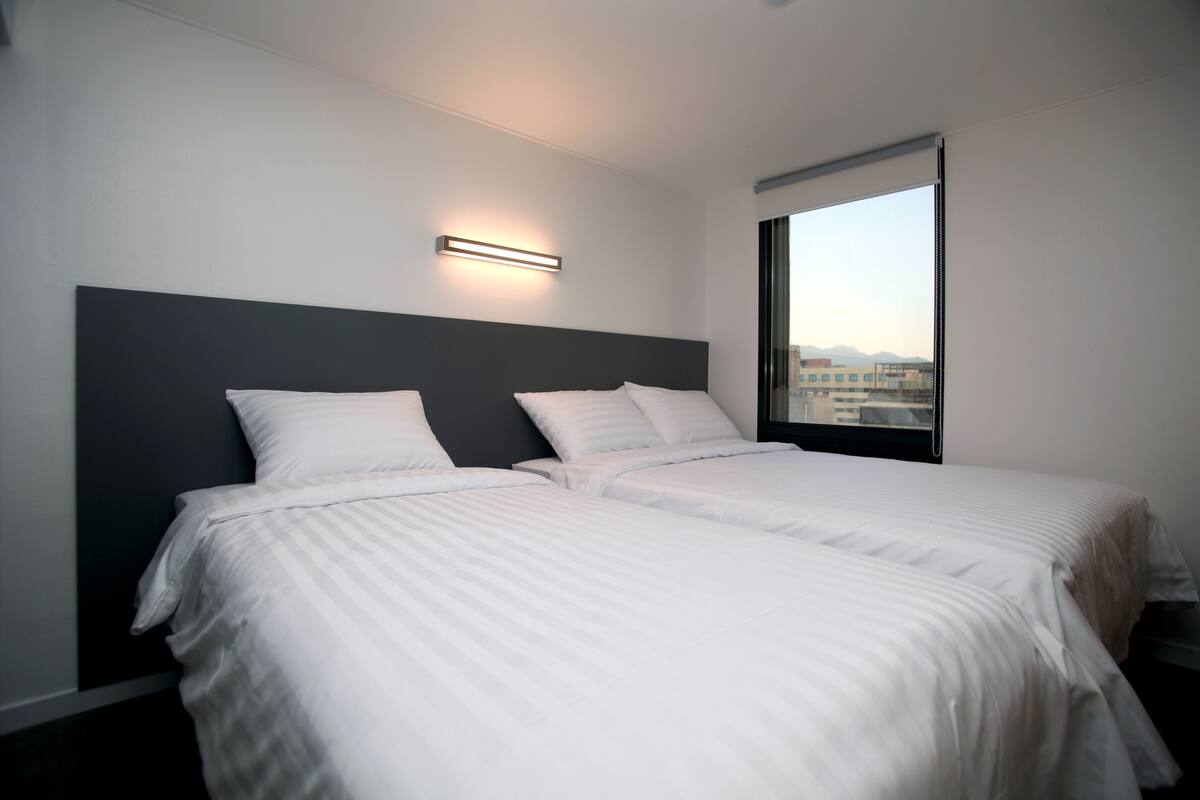 A hotel room with two beds, a modern headboard, a bedside table, and a large window offering a city view.