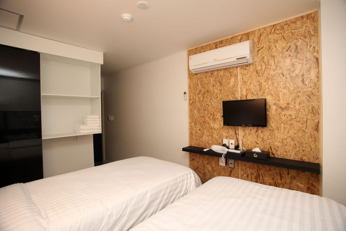 A hotel room with two beds, a mounted TV, an air conditioning unit, and a shelf with toiletries.