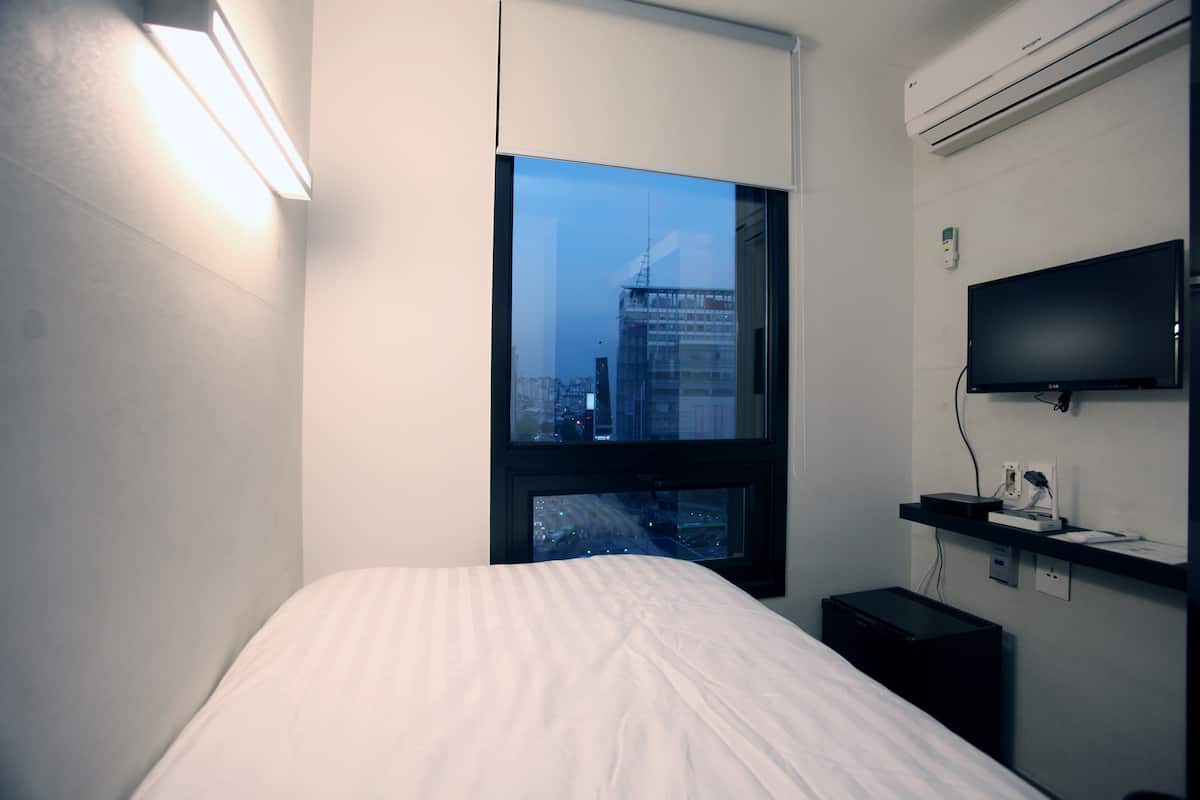 A hotel room with a bed, a TV, a desk, and a window with a city view.