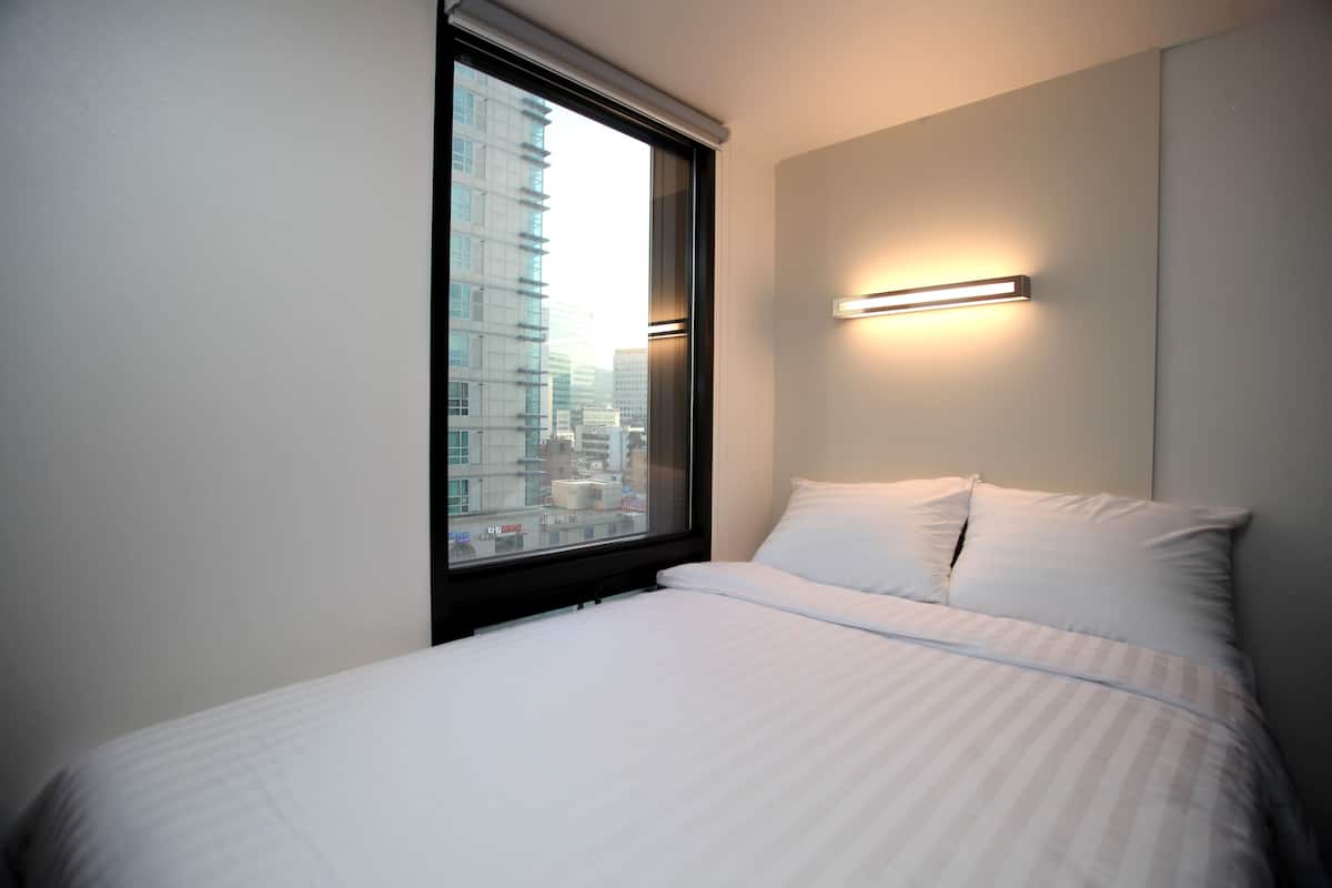 A hotel room with a large window, a bed with white linens, and a wall-mounted light fixture.