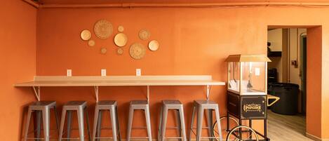 A room with an orange wall, a long table with bar stools, and a popcorn cart.