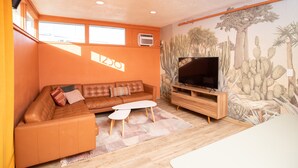 Un salon avec un canapé en cuir marron, une table basse blanche, un téléviseur à écran plat posé sur un meuble en bois, et une fresque murale sur le thème du désert.
