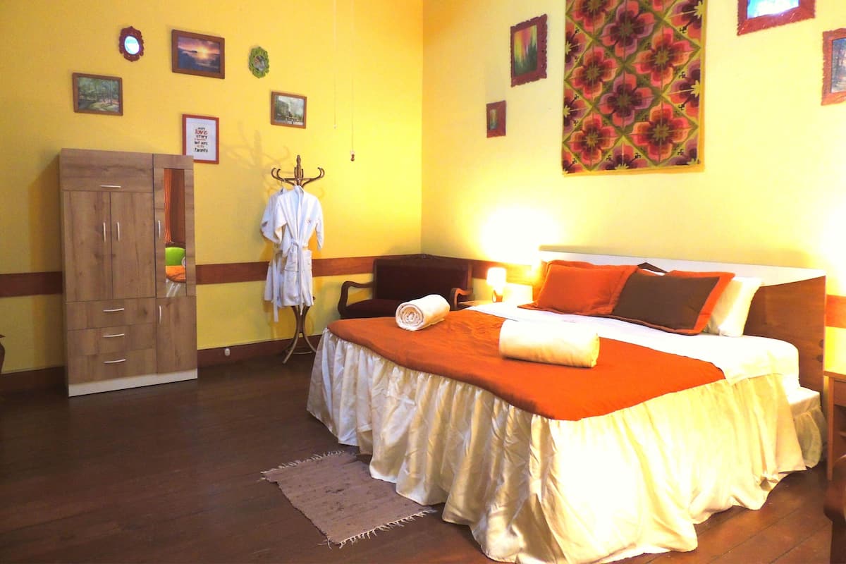 A hotel room with a bed, a wooden wardrobe, a robe on a rack, and a wall with framed pictures.