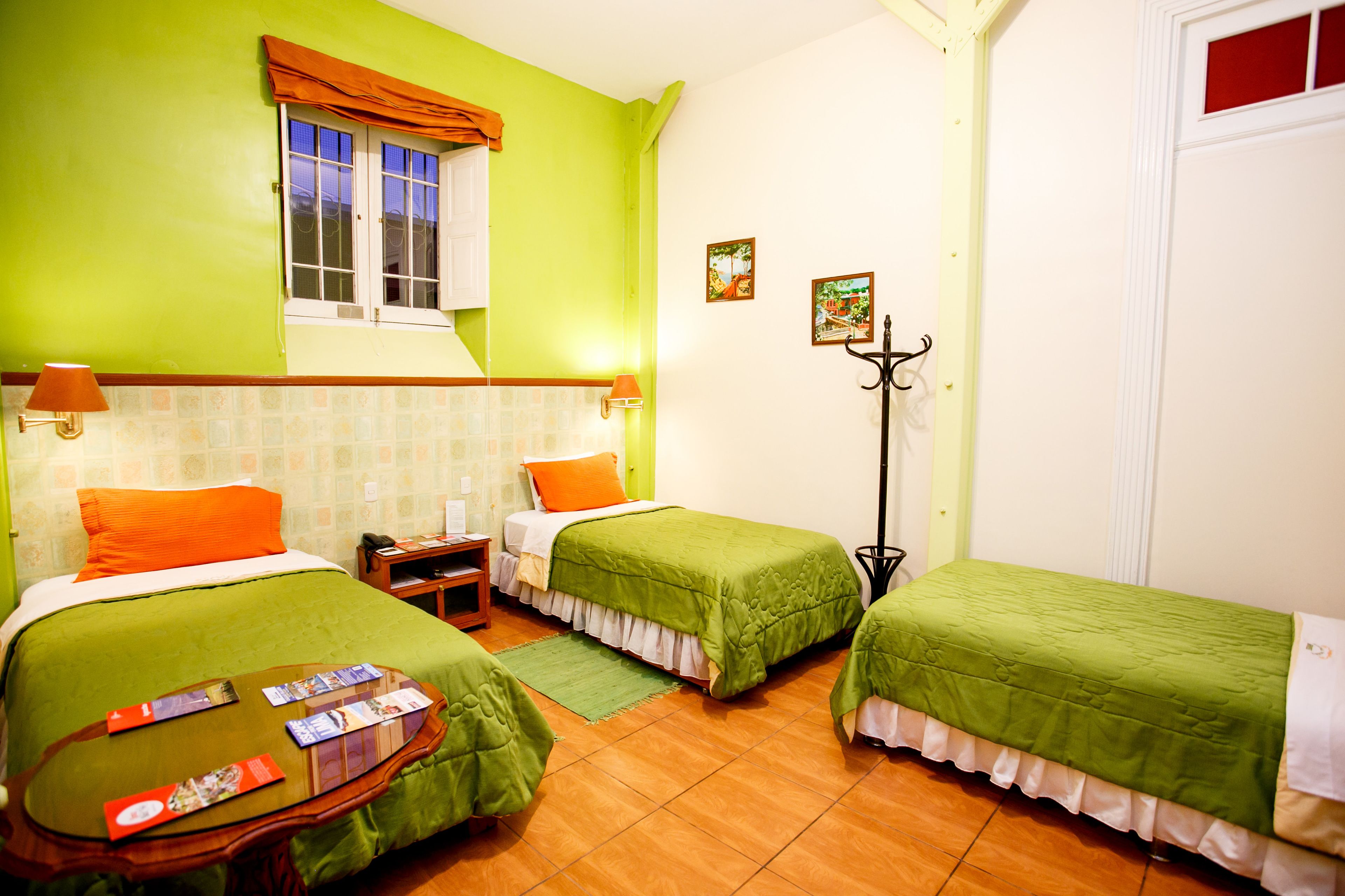 A room with two single beds, a small table with brochures, a window with curtains, and a coat rack.