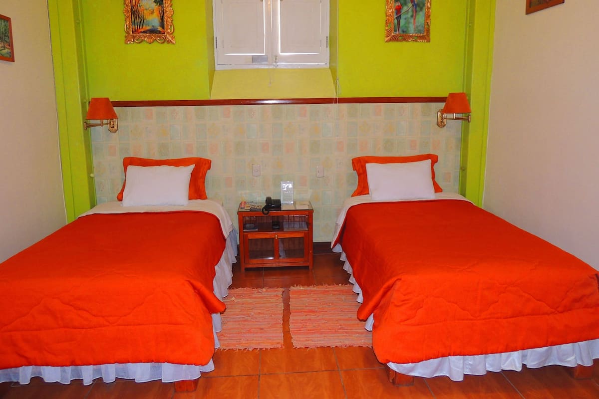 A room with two beds, each with an orange bedspread and white pillows, a wooden nightstand, and a window with white shutters.