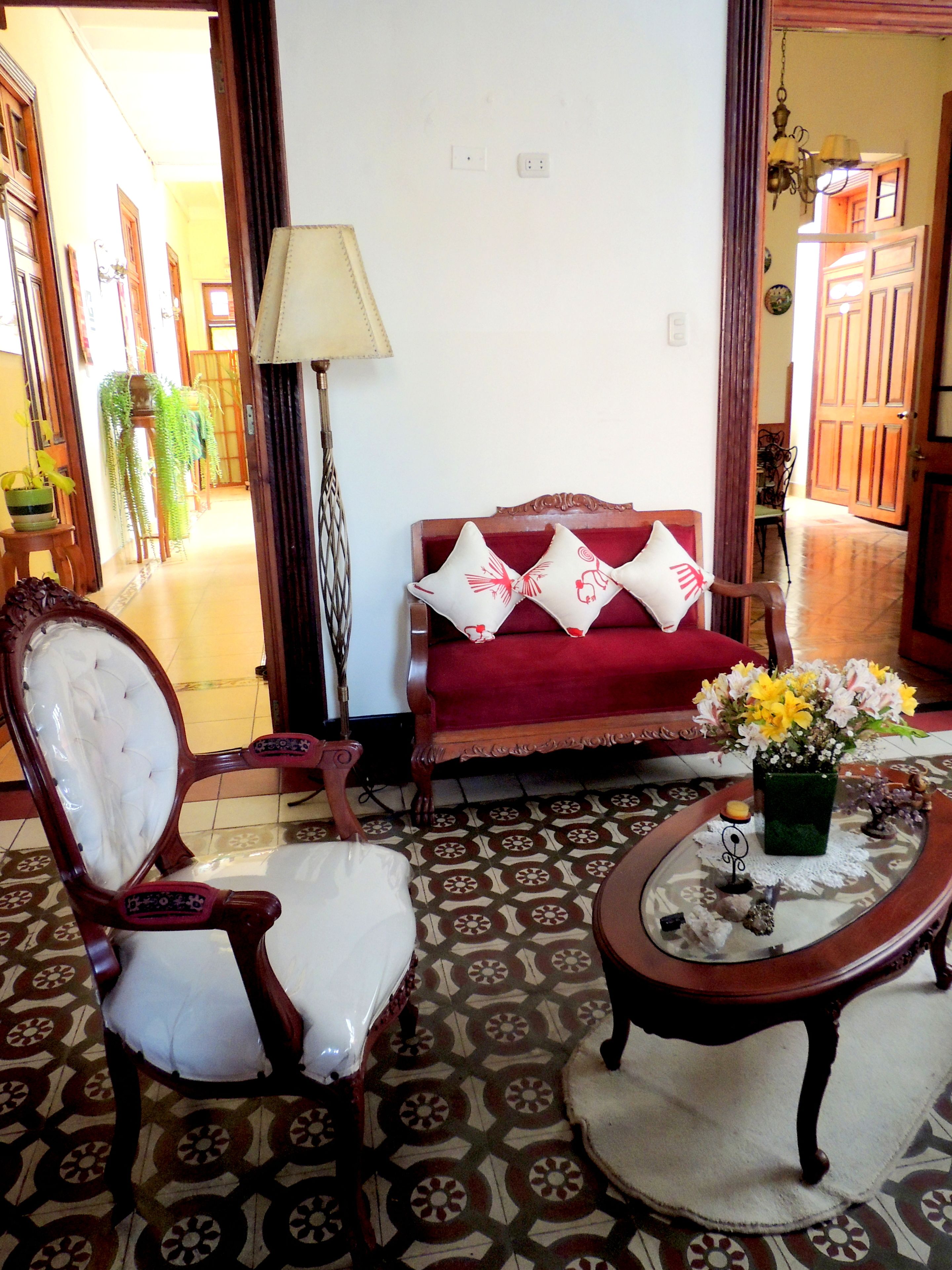 Una sala de estar tradicional con un sofá rojo, una silla de madera, una mesa con un jarrón de flores y baldosas de suelo estampadas.