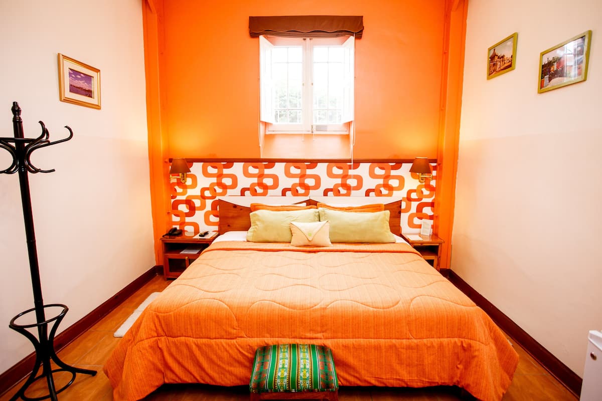 A room with a large bed, a window with curtains, a coat rack, and two framed pictures on the wall.