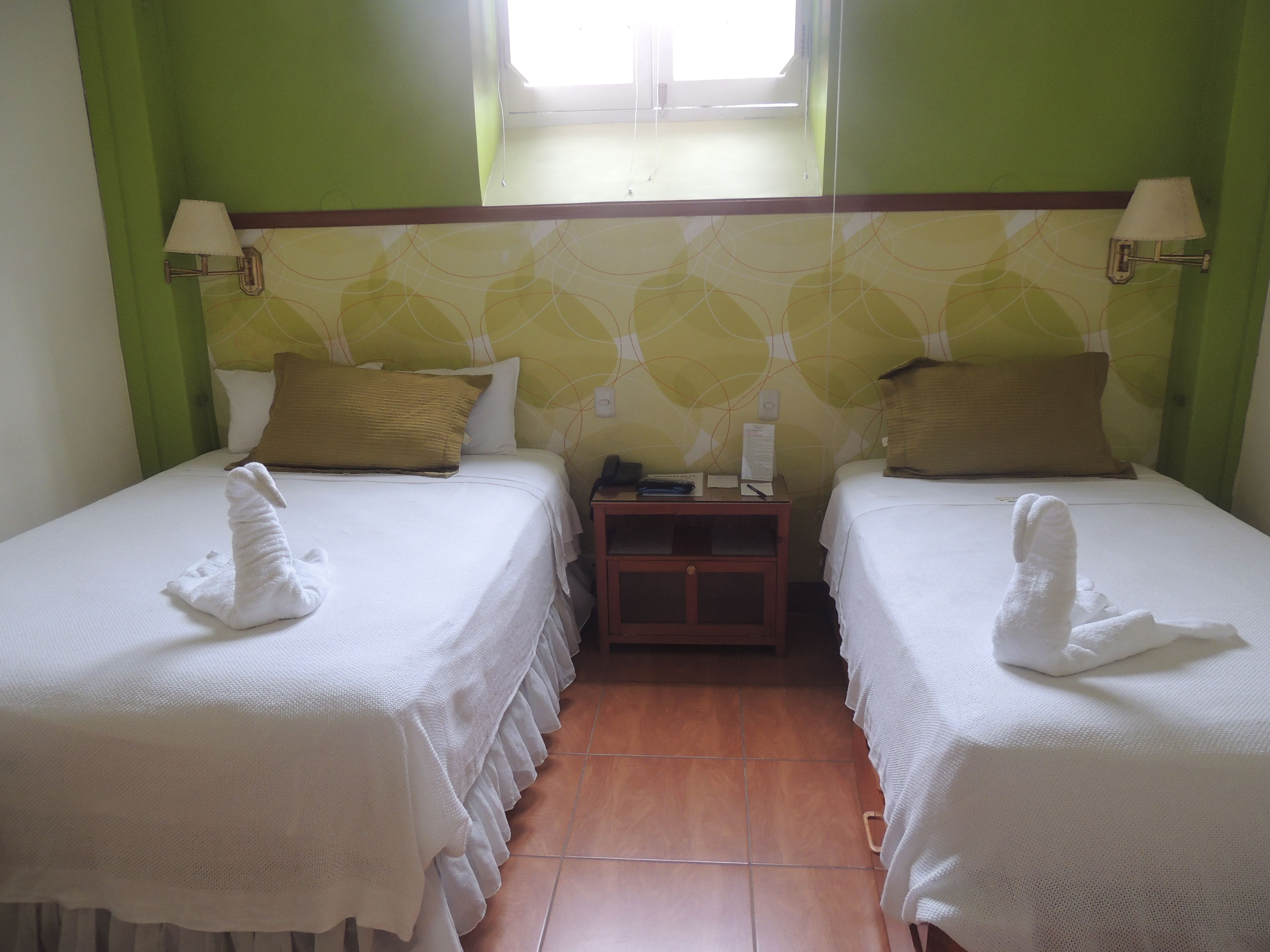 A hotel room with two beds, each with a towel and pillow, a nightstand with a phone and papers, and a window with curtains.