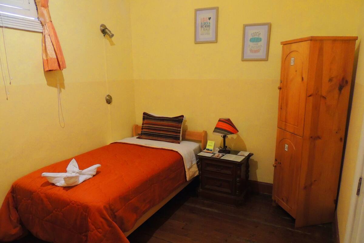 A small, single-bed room with a wooden wardrobe, a bedside table with a lamp, and two framed quotes on the wall.
