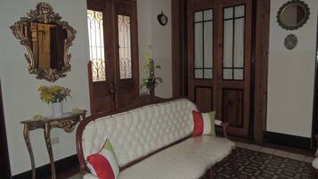 A room with a white cushioned bench, a wooden door with glass panels, a mirror with an ornate frame, and a small table with a vase of flowers.