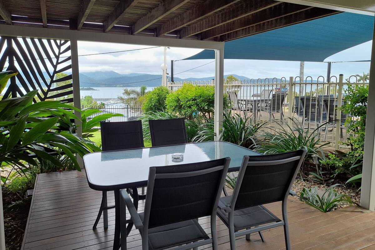 Een overdekt terras met een tafel en stoelen, met uitzicht op een waterpartij en bergen.