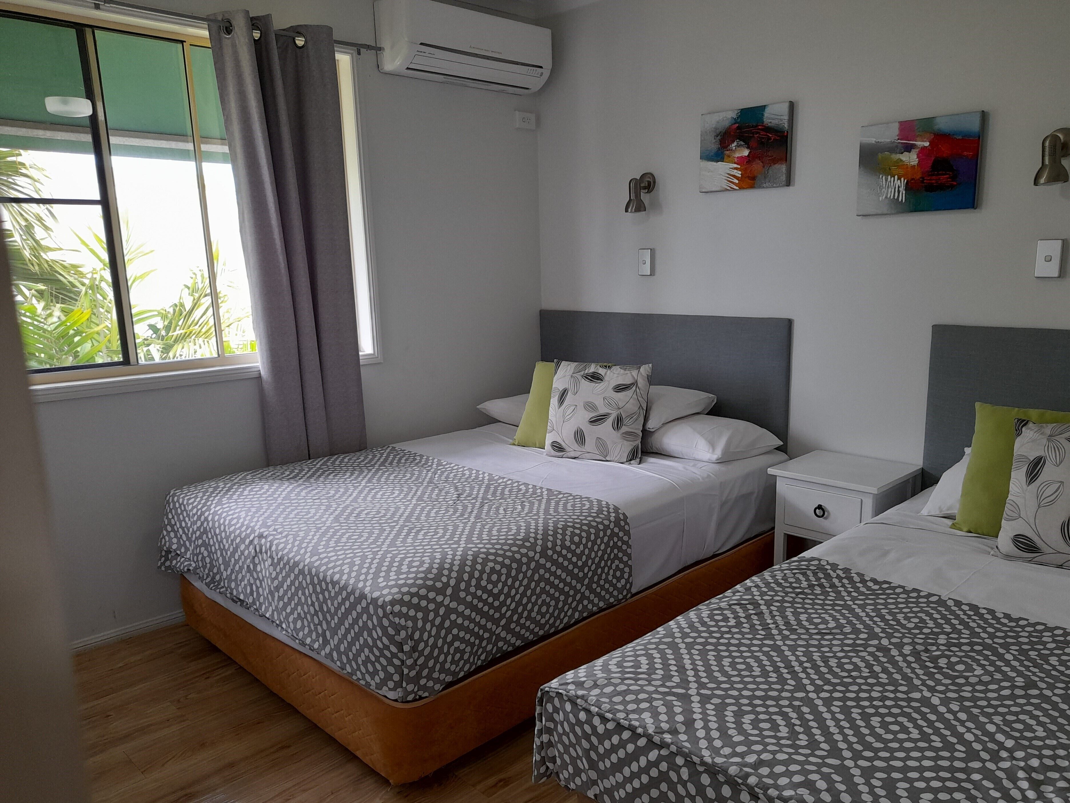 A bedroom with two beds, a window with curtains, an air conditioner, and a wall with two paintings.