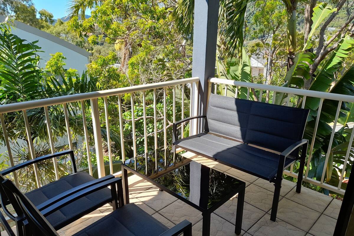 Een balkon met twee zwarte stoelen en een tafel, omgeven door een witte balustrade, met uitzicht op een weelderige tuin en een bergachtig landschap.
