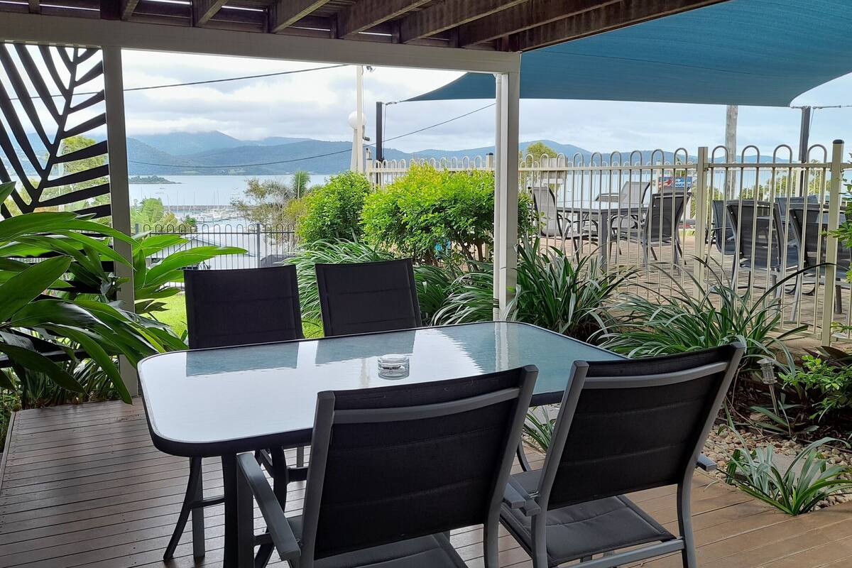 Een terras buiten met een glazen tafel en stoelen, omgeven door planten en uitzicht op zee.