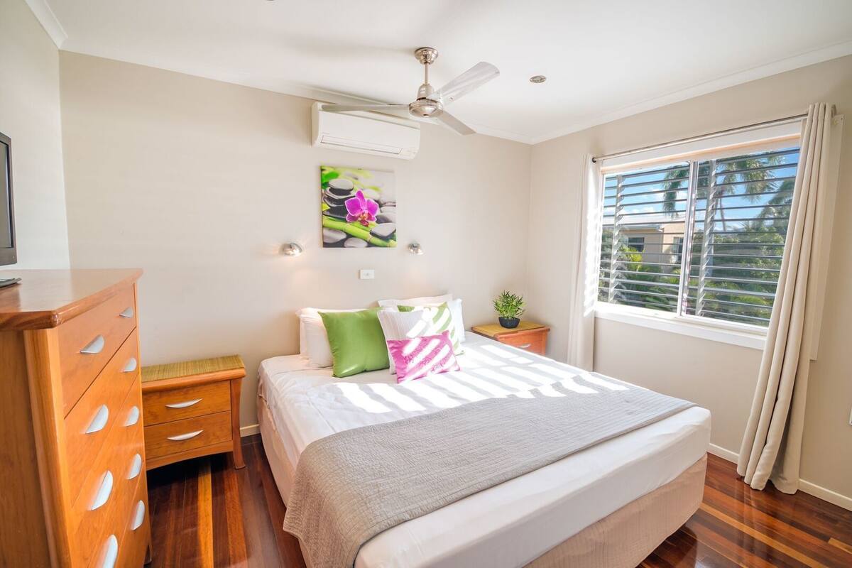 Een slaapkamer met een bed, nachtkastje, dressoir, plafondventilator, raam met gordijnen en een wandairconditioner.