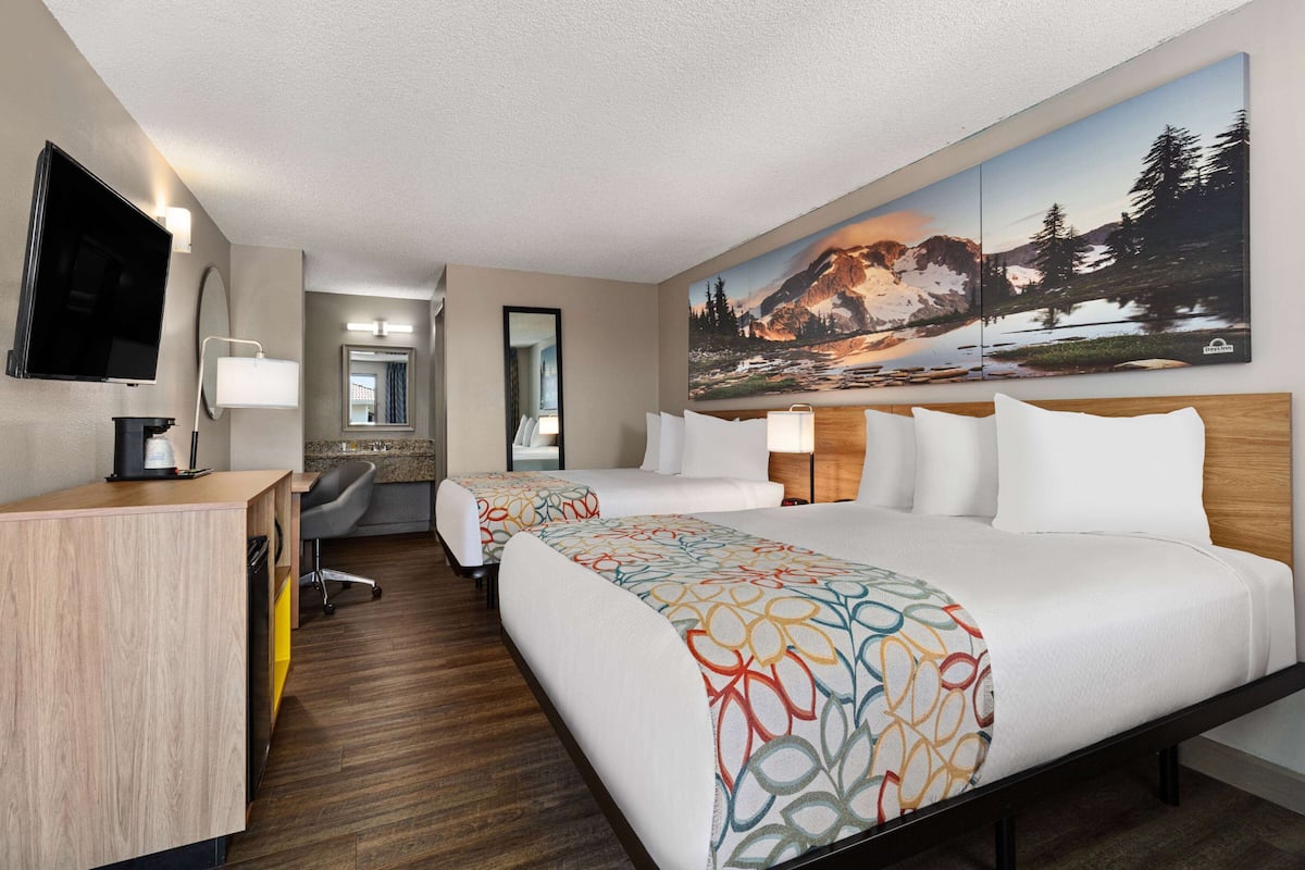 Habitación de hotel con dos camas, un escritorio, un televisor y un cuadro grande de un paisaje montañoso en la pared.