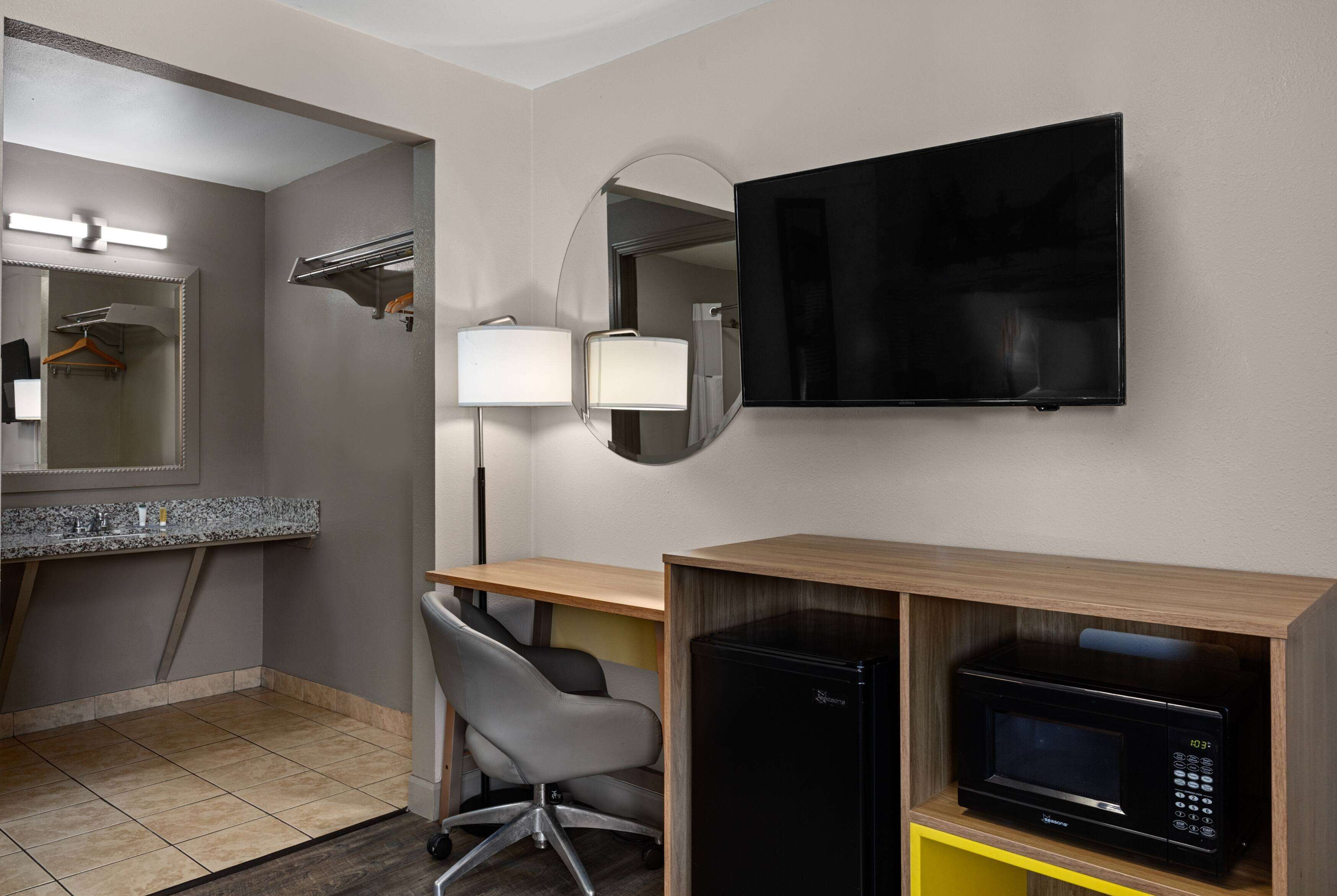 A hotel room with a bathroom, a desk, a chair, a TV, and a microwave.