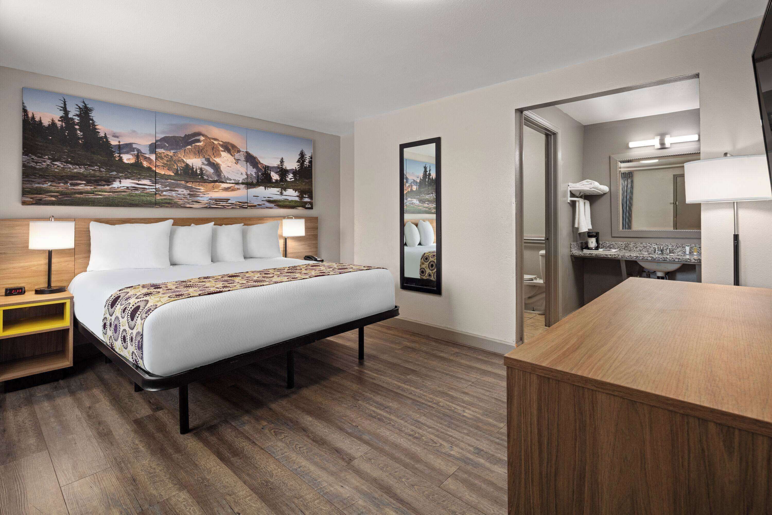 A hotel room with a large bed, bedside tables, a wooden desk, a bathroom with a mirror, and a mountain landscape painting on the wall.