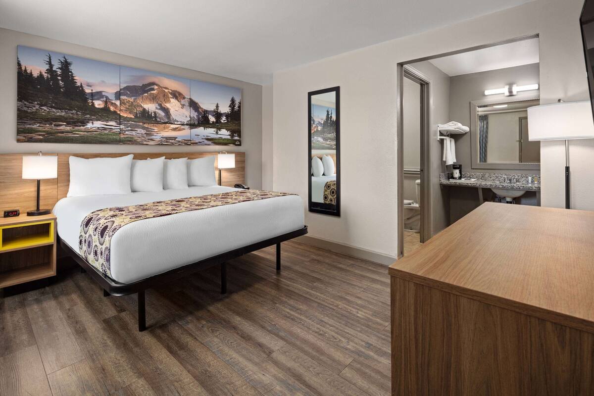 Habitación de hotel con una cama grande, una mesita de noche con lámpara, un baño con lavabo y espejo, y piso de madera.
