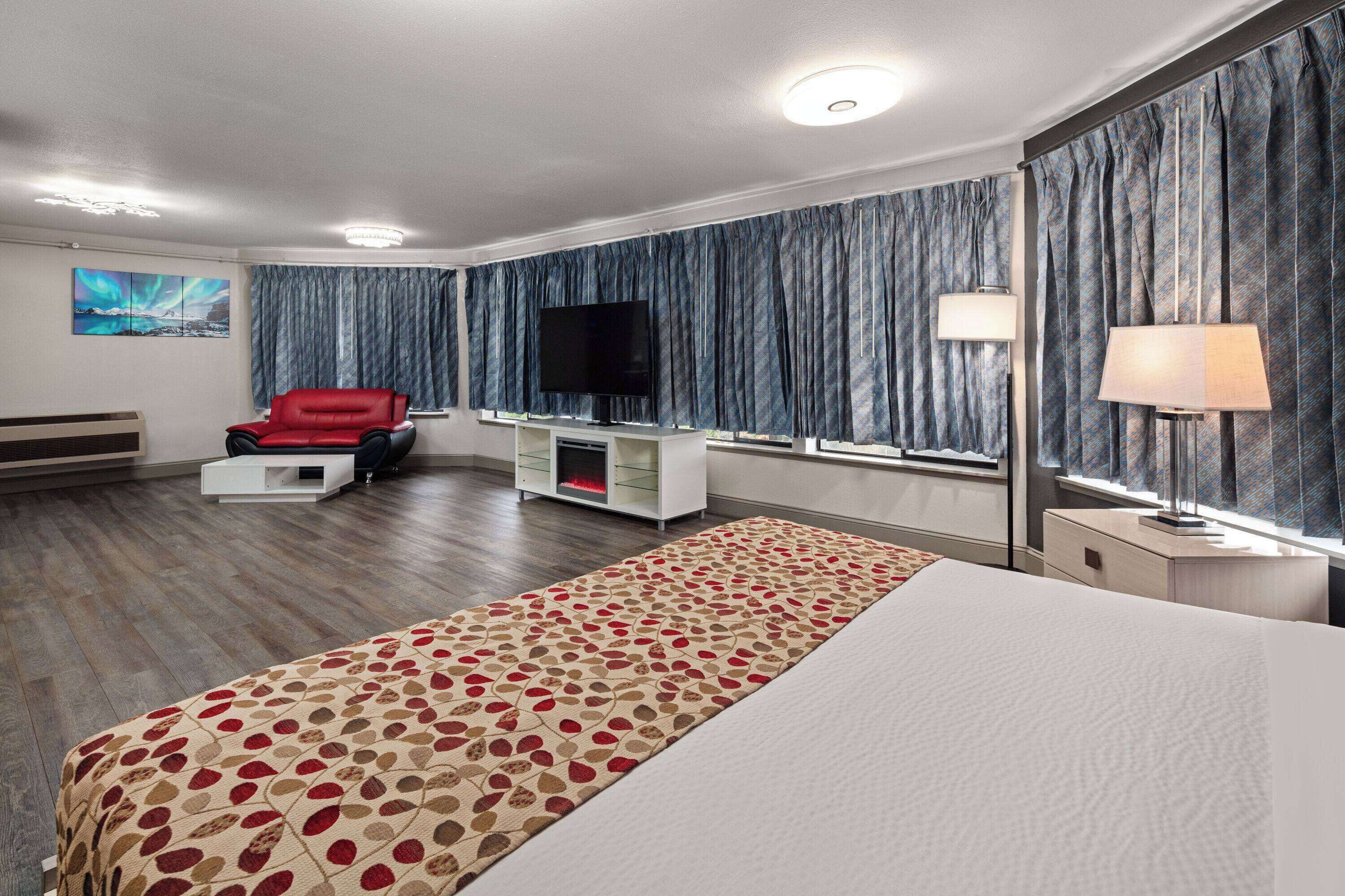 Habitación de hotel con una cama grande, una silla roja, un televisor de pantalla plana, un mueble para televisión, una lámpara y una ventana con cortinas.