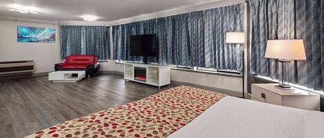 Habitación de hotel con una cama grande, una silla roja, un televisor de pantalla plana, un mueble para televisión, una lámpara y una ventana con cortinas.