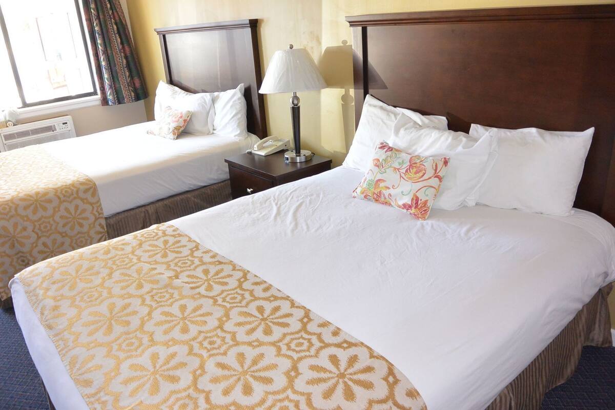 Habitación de hotel con una cama grande, dos camas más pequeñas, una mesita de noche con lámpara y una ventana con cortinas.