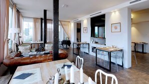 A modern living room with a leather sofa, dining table, and chairs. There is a kitchen area with a large window and a flat-screen TV.