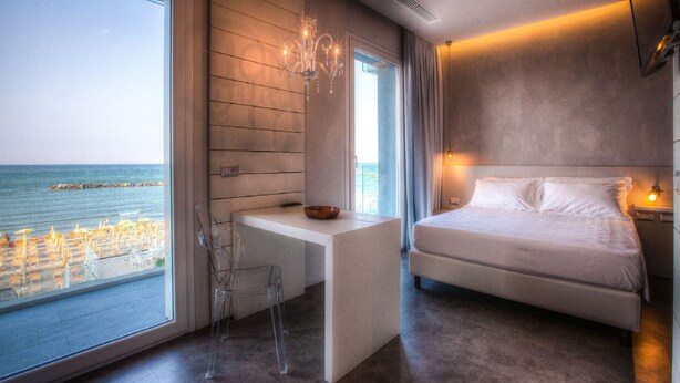 A modern hotel room with a large window overlooking the sea, a bed, a desk, and a clear chair.