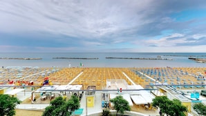 A beach with rows of sun loungers and umbrellas, a pool area with tables and chairs, and a view of the sea and distant boats.