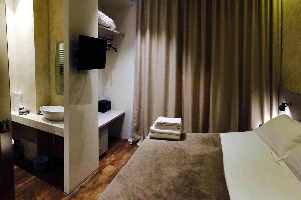 Une chambre d’hôtel avec un lit, une table de chevet, un miroir, un téléviseur fixé au mur et un rideau.