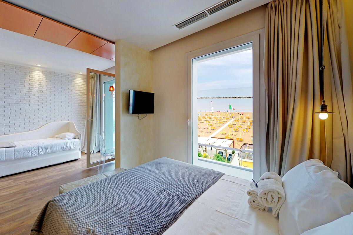 Une chambre d’hôtel moderne avec un mur en briques blanches, un grand lit recouvert d’une couverture à motifs gris et blancs, un téléviseur à écran plat fixé au mur et une vue sur une plage avec des parasols, visible à travers une grande fenêtre.