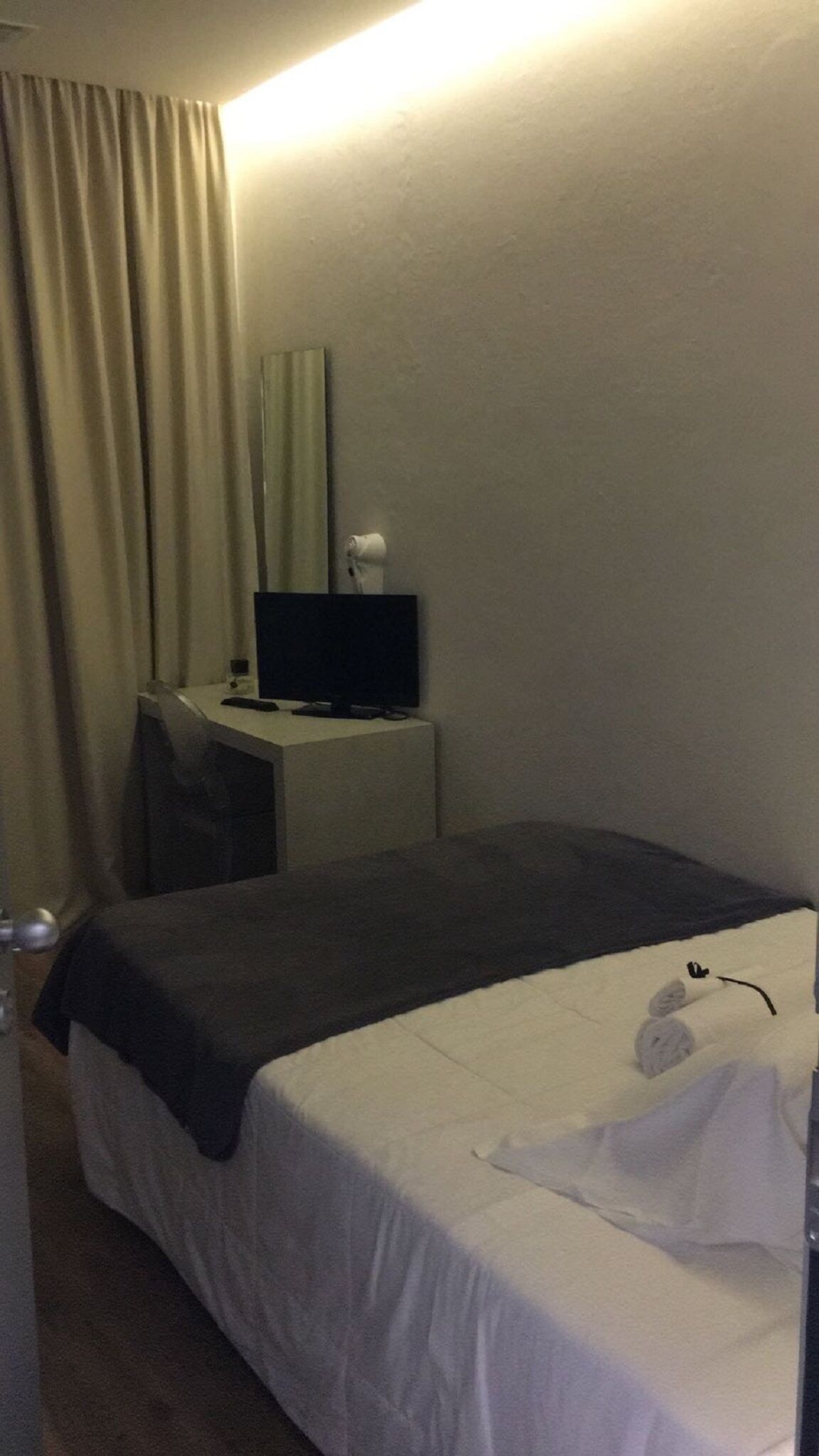 Hotellihuone, jossa on sänky, työpöytä tietokoneella, tuoli ja ikkuna, jossa on verhot.
