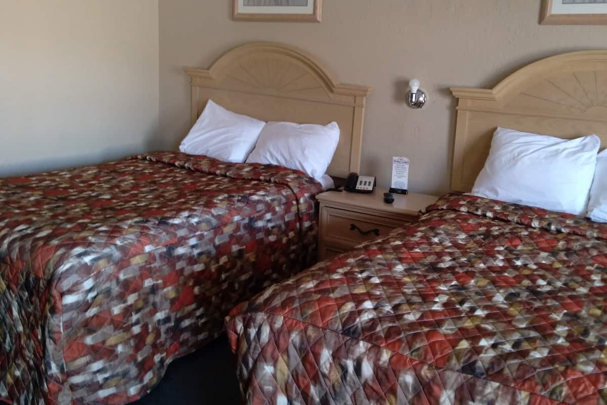 A hotel room with two beds, each with a patterned bedspread, a wooden headboard, a nightstand, and a wall-mounted lamp.