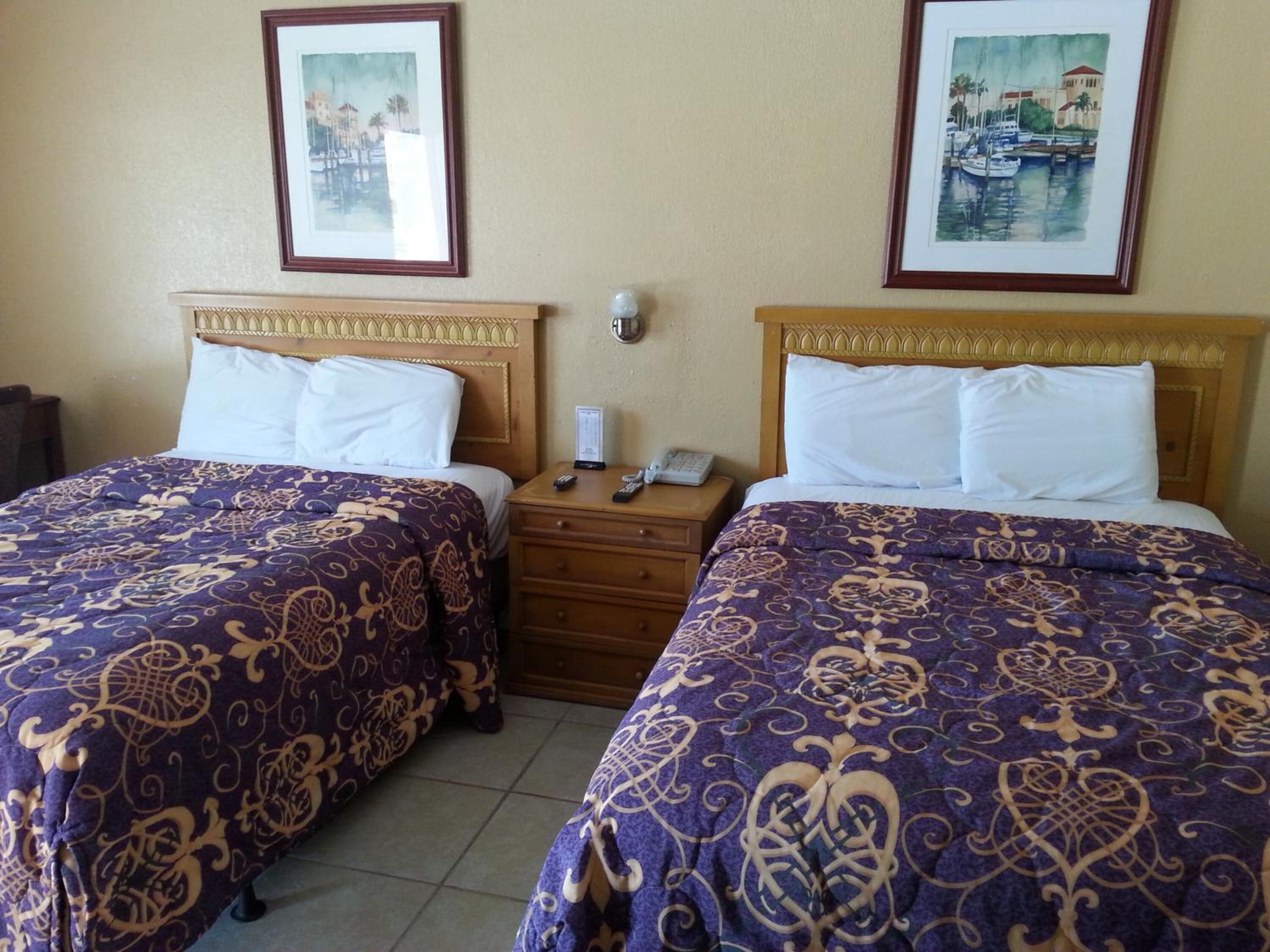 A hotel room with two beds, each with a purple and gold patterned bedspread, wooden headboards, and white pillows. There are framed pictures on the walls, a nightstand with a phone and remote controls, and a small drawer.