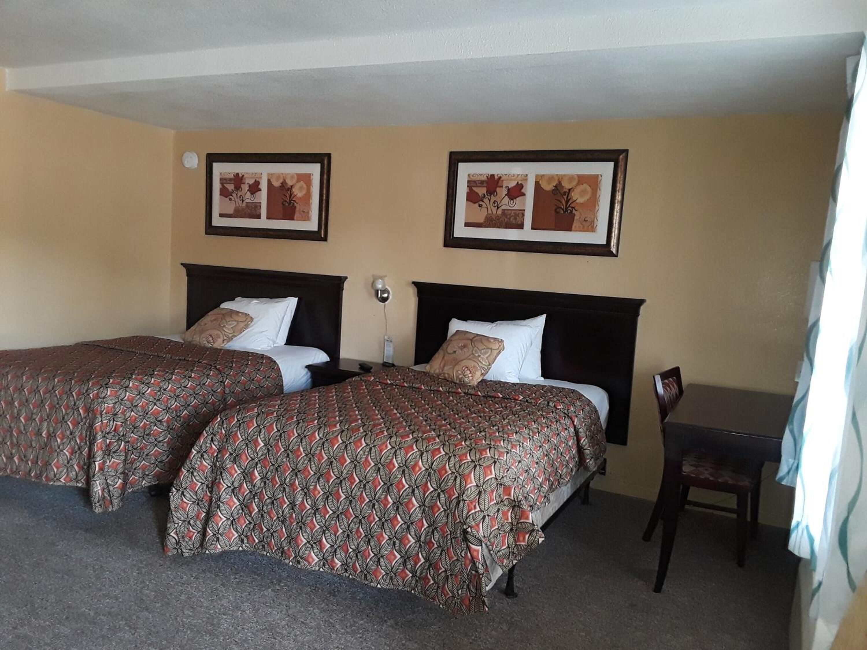 A hotel room with two beds, each with patterned bedspreads, a desk, and two framed pictures on the wall.