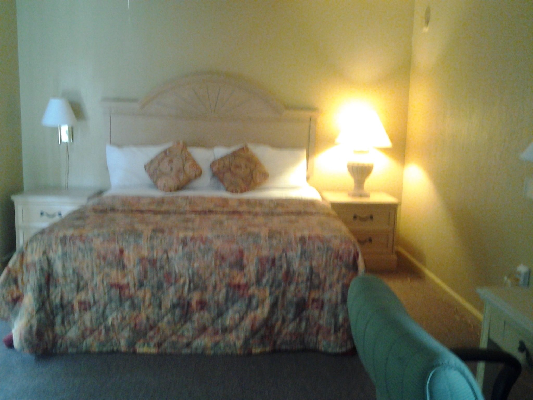 A bedroom with a bed, two pillows, a nightstand with a lamp, and a chair.