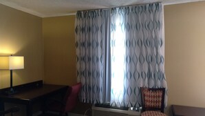 A hotel room with a desk, a chair, a small table, and a window with patterned curtains. - Budget Lodge of Mount Dora (Mount Dora)