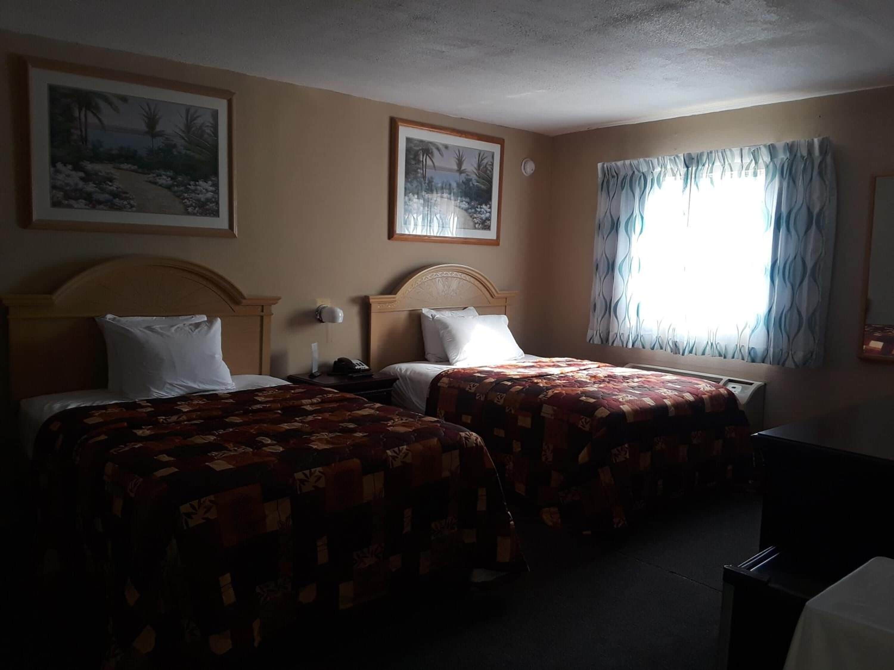 A hotel room with two beds, a nightstand, a lamp, and a window with curtains.